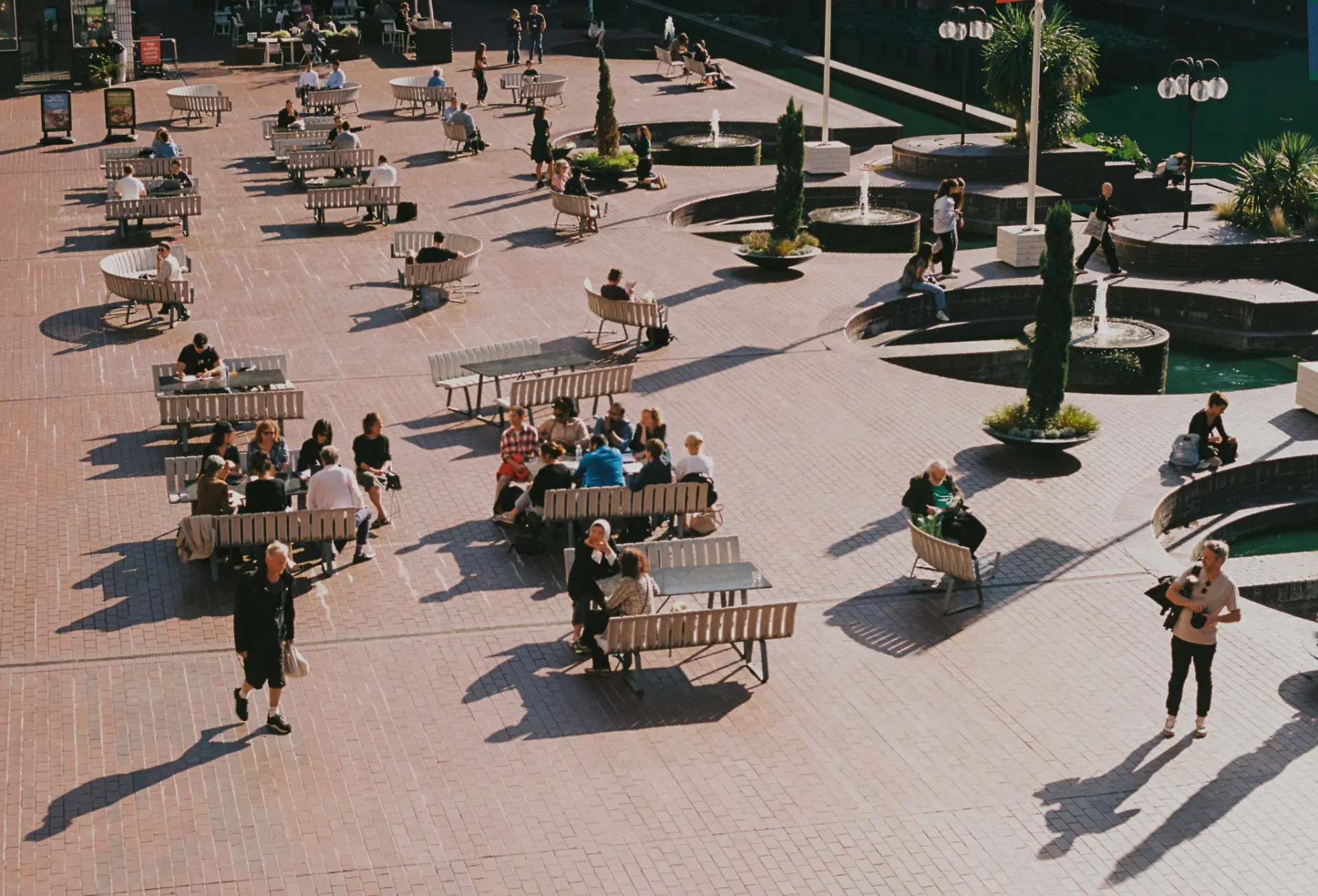People gathering among benches and fountains in a sunlit public plaza